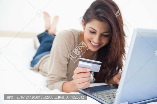面帶微笑的女人躺在床上，手里拿著信用卡，手里拿著筆記本；雙腿交叉。