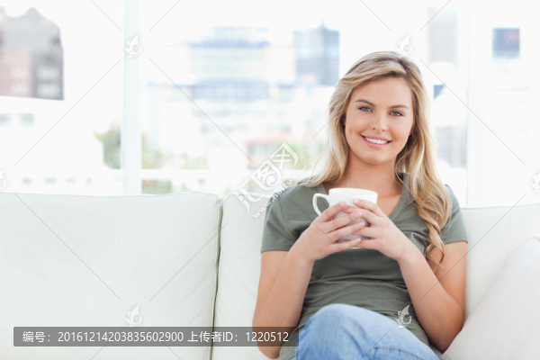 一個(gè)女人在肩前舉著一個(gè)杯子，她坐在沙發(fā)上微笑著。