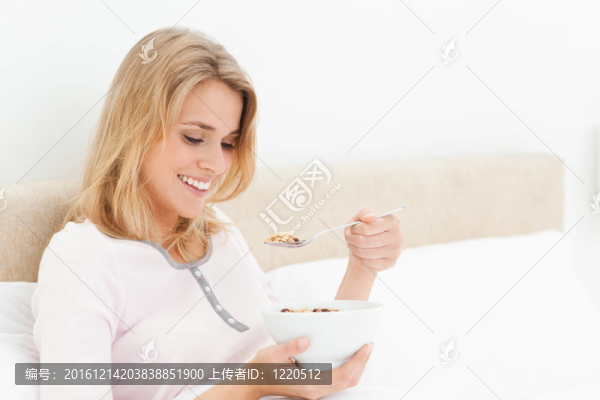 一個(gè)女人坐在床上，手里拿著一個(gè)碗，一邊拿著勺子，一邊看著勺子和微笑。