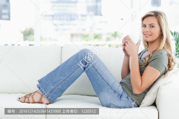 一個女人側(cè)身坐在沙發(fā)上；腿微微抬起；微笑著，一個杯子舉到嘴邊。