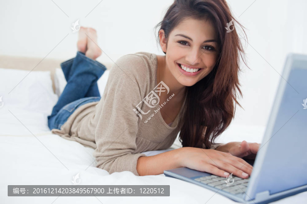 一個(gè)面帶微笑的女人在床前躺在筆記本電腦前微微抬起腿。