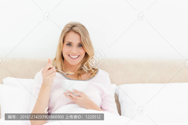 一個女人坐在床上，手里拿著一碗麥片粥，手里拿著一把麥片粥，一邊向前看，一邊微笑著。