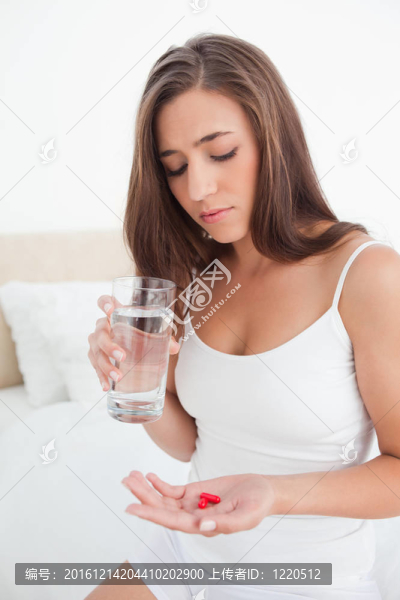 一個女人低頭看著手里拿著的兩個紅色藥丸，手里拿著一杯水。