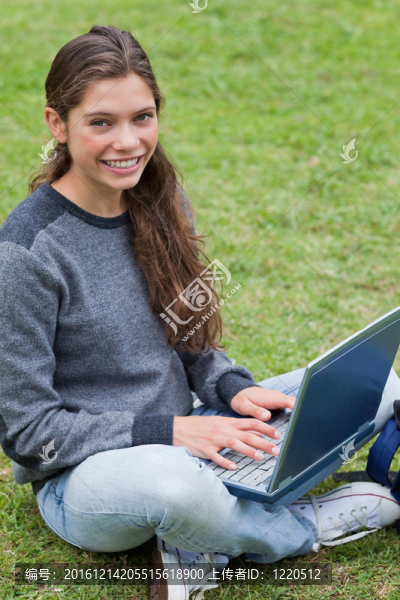 微笑的年輕女孩坐在公園的草地上，在筆記本電腦上打字