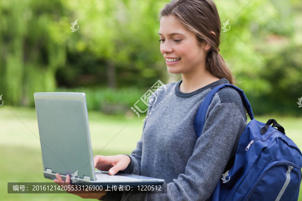 年輕的面帶微笑的女人拿著她的筆記本電腦，站在一個(gè)帶著頭發(fā)的公園里