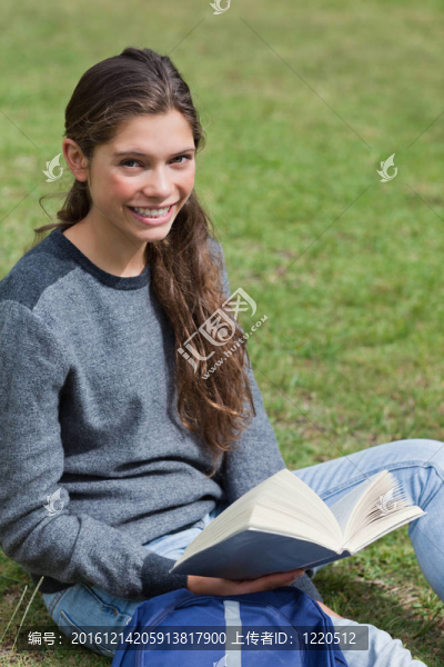 微笑的年輕女孩在公園的草地上坐著，手里拿著一本打開的書