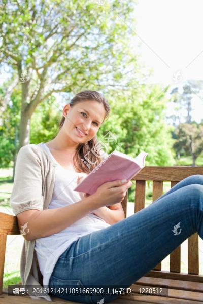 微笑的女人坐在公園的長椅上，拿著一本書