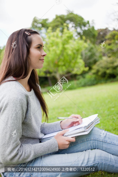 年輕而放松的女孩看著遠(yuǎn)處，坐在公園里拿著一支筆和一本筆記本