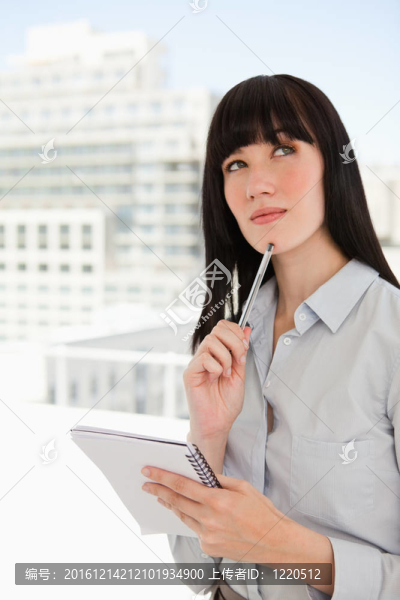 一個(gè)女人在辦公室里拿著一個(gè)記事本和一支筆放在下巴上，她想