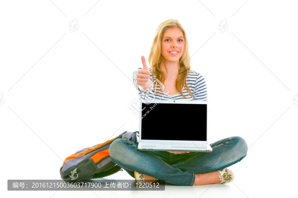 微笑的漂亮女孩坐在地板上用筆記本電腦，并顯示拇指孤立的白色