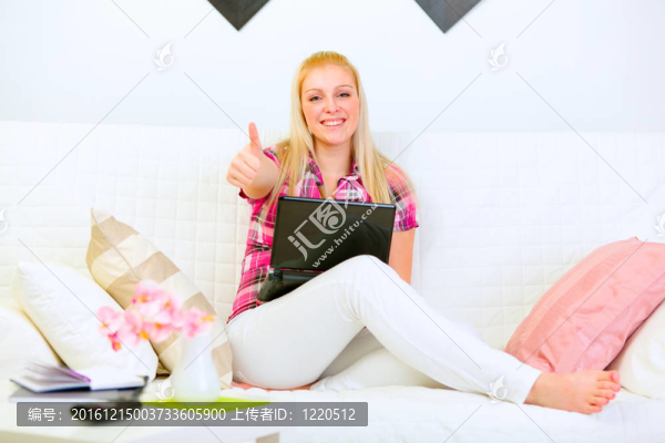 微笑的漂亮女人坐在沙發(fā)上，用筆記本電腦，豎起大拇指