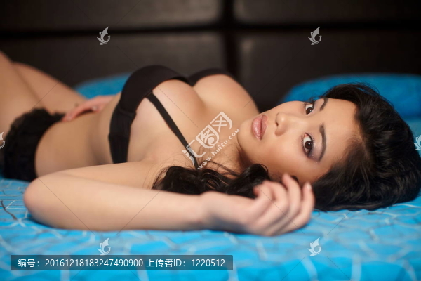 年輕性感的黑發(fā)中國(guó)魅力女人模型與黑色內(nèi)衣長(zhǎng)而卷曲的頭發(fā)躺在床上的藍(lán)色明亮的室內(nèi)