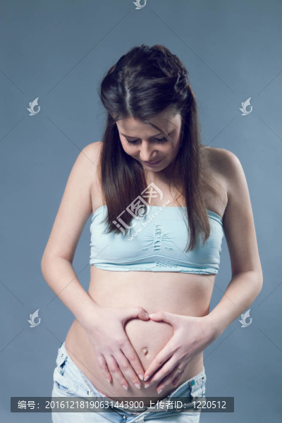 美麗的孕婦白女人wearinglight藍(lán)色上衣和牛仔褲。長黑發(fā)。雙手放在肚子上；手指在肚臍上方呈心