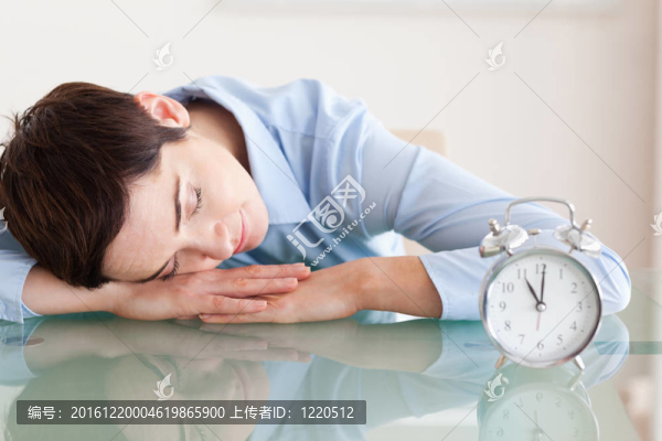 沉睡的可愛的黑發(fā)女人把頭靠在桌子旁邊的辦公室里的鬧鐘