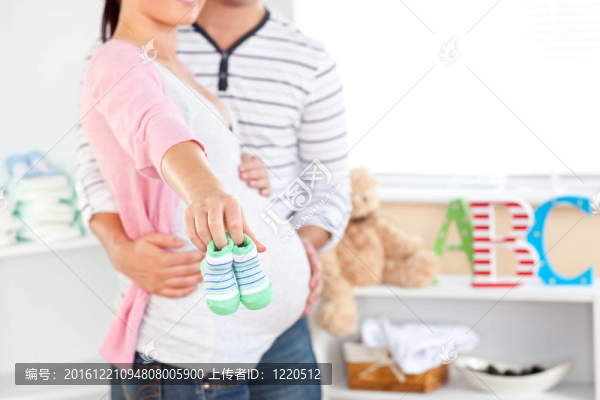 一個明亮的孕婦抱著嬰兒鞋，而丈夫接觸她的肚子在未來的嬰兒在家里的房間