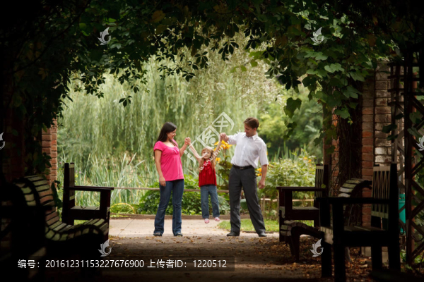小女孩穿著紅色連衣裙和爸爸媽媽在公園里的植物隧道里。女孩在父母的手上搖動(dòng)。橫向格式