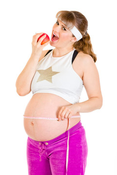 微笑美丽的孕妇抱着卷尺，吃着白色的苹果