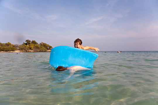 男孩是在一个美丽的海边的一个小的冲浪板与清澈的水和蓝天冲浪；他的兄弟拥有冲浪板的背