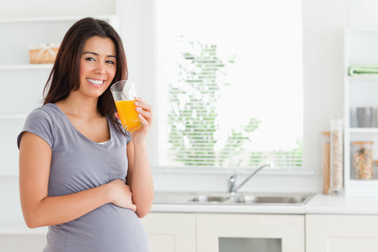 美丽的孕妇站在厨房里喝着一杯橙汁